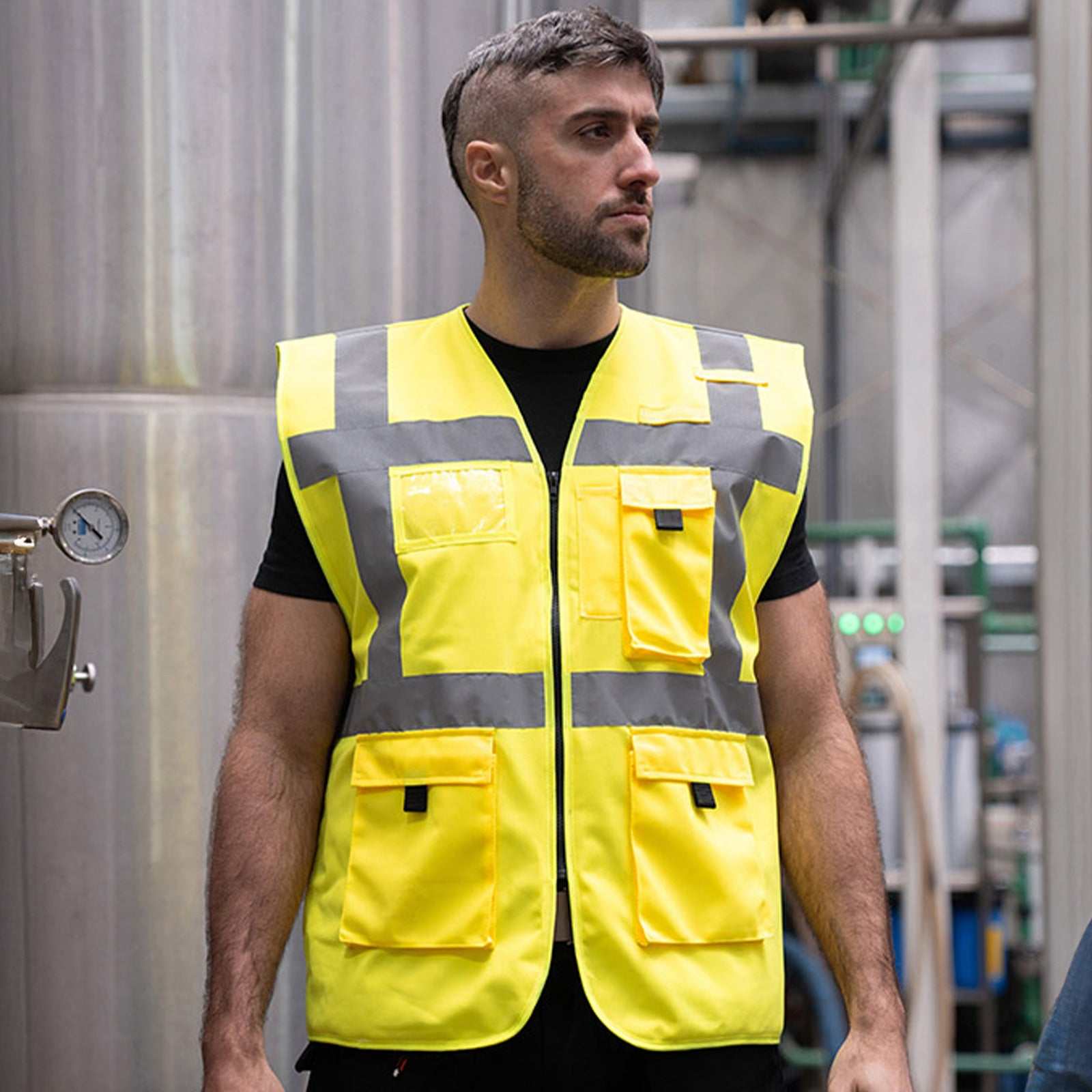 Mann trägt gefütterte Warnweste Comfort-Fit „Wismar“ in Gelb mit reflektierenden Streifen in industrieller Umgebung